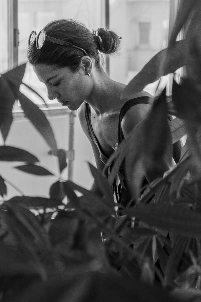 Vilelmini standing behind plants, softly lit, focused in a quiet moment of preparation.